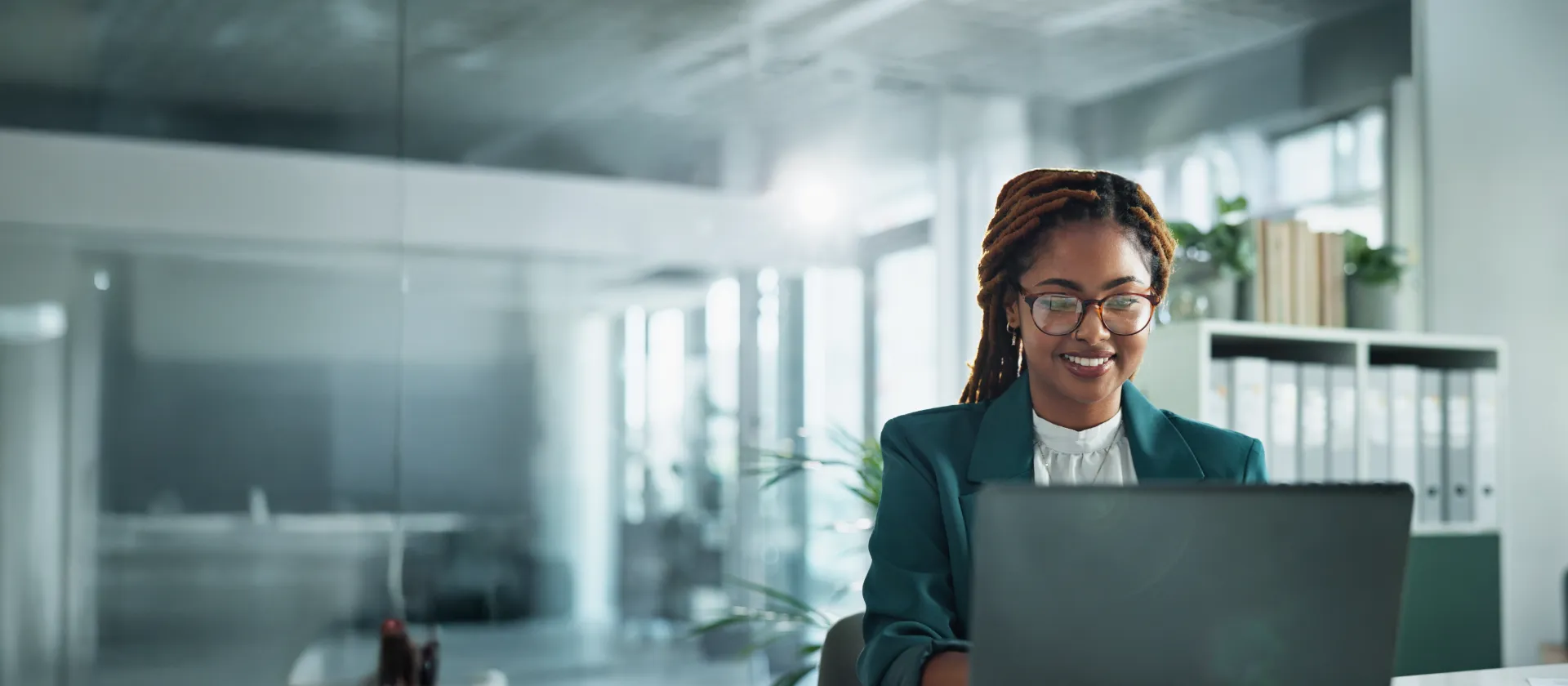 Businesswoman with glasses smiling while working on a laptop in a modern office.